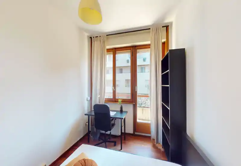 Camera da letto luminosa e minimale con scrivania vicino alla finestra, vista parziale del letto in primo piano e una libreria alta — ideale per una persona o uno studente.