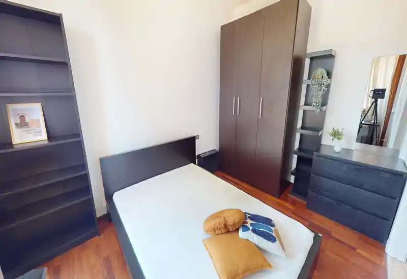 Camera da letto luminosa e arredata con semplicità, letto singolo, armadio e scaffali in legno scuro e comò con specchio. Linee pulite e pavimento in legno caldo rendono l'ambiente ordinato e accogliente, ideale per una persona.