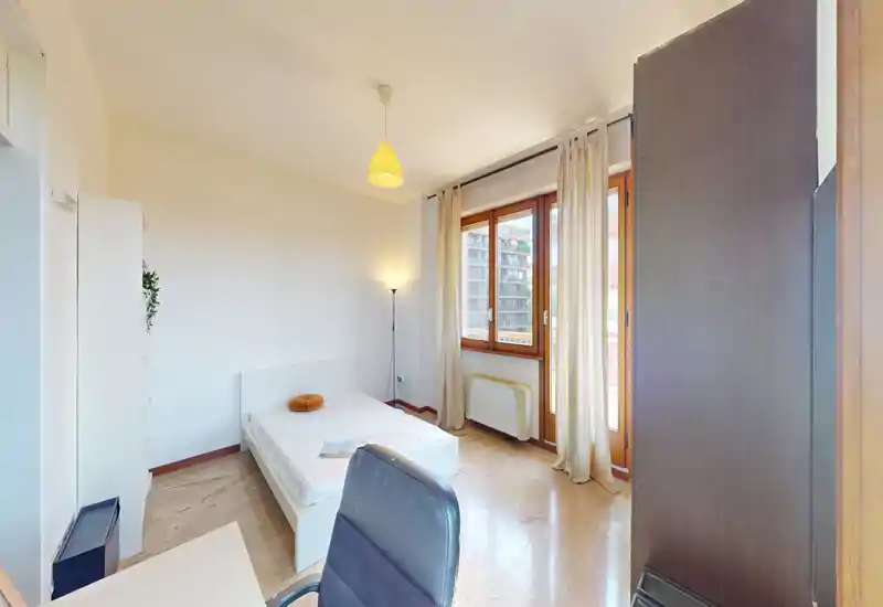 Camera da letto luminosa e arredata con semplicità con letto matrimoniale, ampia finestra e angolo scrivania; toni neutri e luce naturale creano uno spazio arioso e funzionale, ideale per affitti brevi.