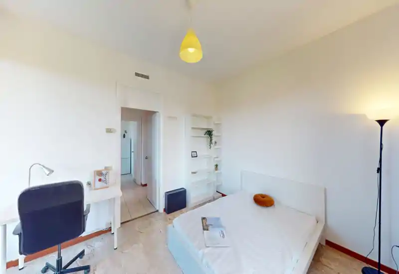 Camera da letto luminosa e minimale con letto matrimoniale, lampada e piccolo angolo studio. Pareti bianche e mensole semplici creano un ambiente arioso e ordinato.