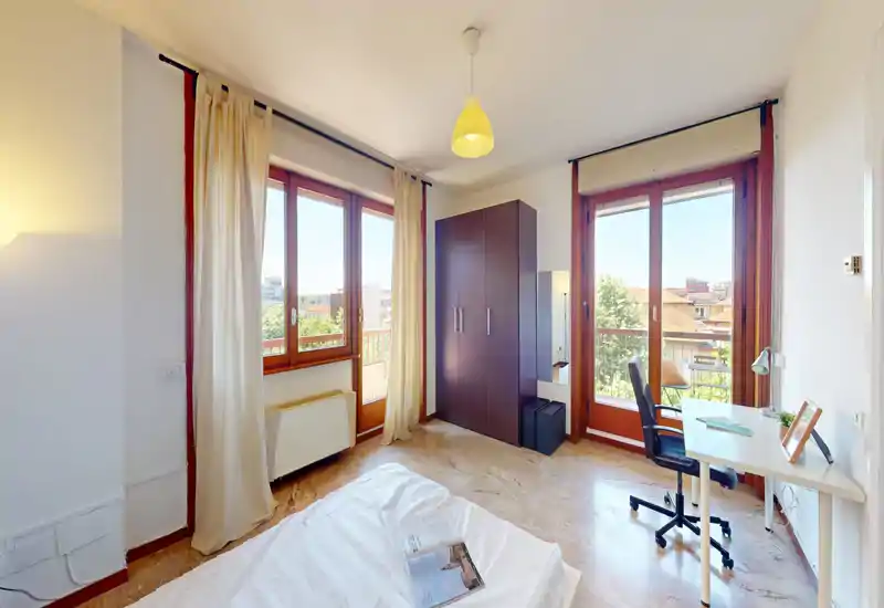 Camera da letto luminosa e ariosa con ampie finestre e un letto matrimoniale parzialmente visibile in primo piano. Un armadio e una scrivania con sedia si affacciano sul balcone, offrendo molta luce naturale e un'area lavoro funzionale.