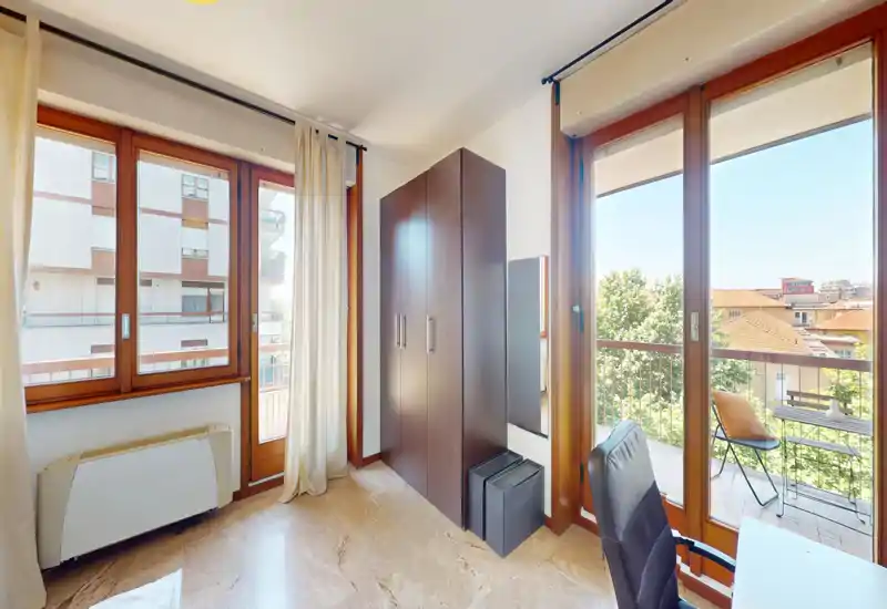 Bright bedroom corner with large windows and balcony access, featuring a wardrobe, desk chair and curtains. The space receives abundant natural light and offers a small balcony with city views.