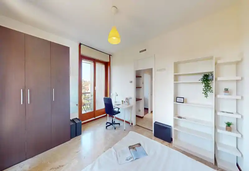 Camera da letto luminosa e ordinata con letto matrimoniale in primo piano, armadio a muro, scrivania vicino alla porta del balcone e scaffalatura con piante.