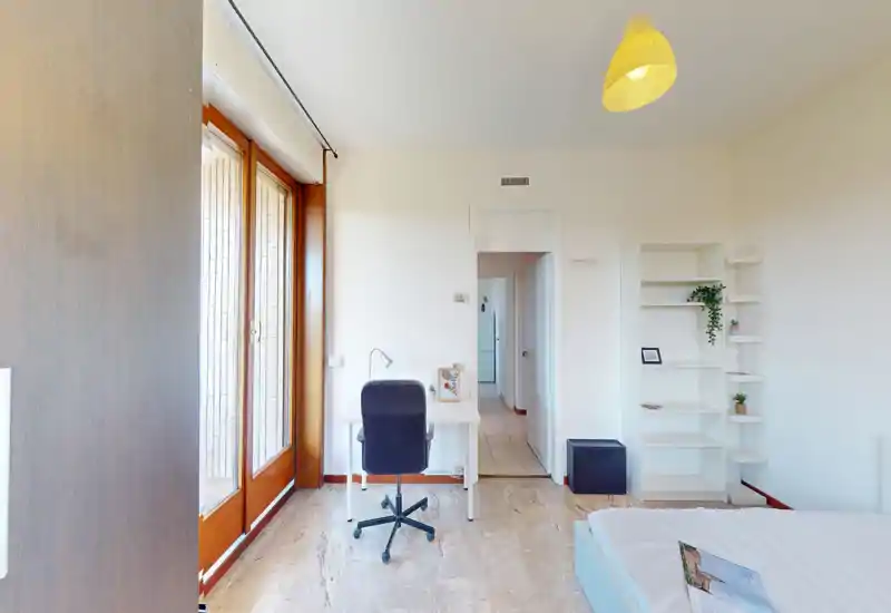 Bright bedroom with a single bed, desk and shelving; large glass doors provide abundant natural light and a pleasant, airy atmosphere.
