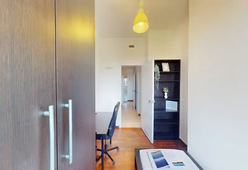 Camera compatta con letto singolo, armadio a muro, scrivania e scaffale; corridoio luminoso che conduce ad altre stanze. Layout pulito e funzionale, ideale per studenti o soggiorni brevi.