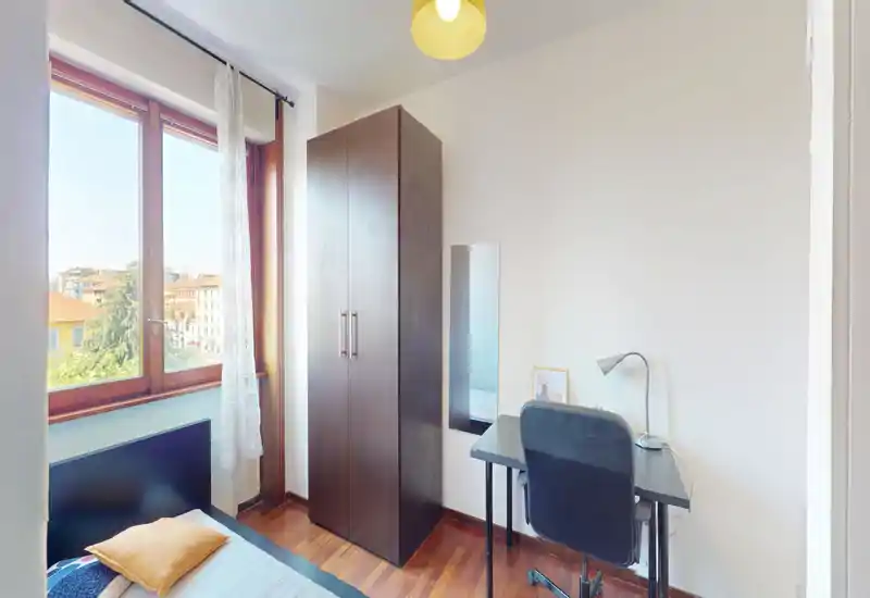 Camera da letto piccola e luminosa con letto singolo, armadio alto e scrivania accanto a una grande finestra che garantisce luce naturale. Pavimento in legno e arredo semplice e moderno rendono lo spazio ordinato e funzionale, ideale per uno studente o una persona sola.