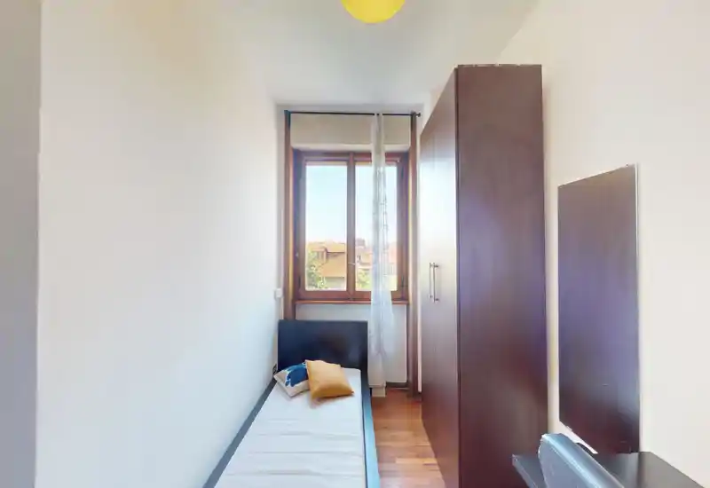 Camera singola compatta con disposizione stretta, pavimento in legno e armadio; la luce naturale dalla finestra rende l’ambiente luminoso e accogliente.