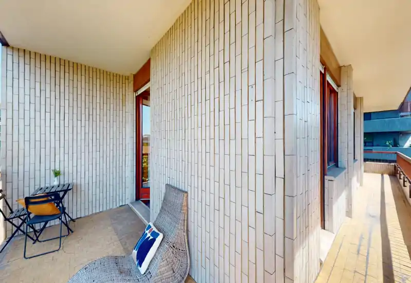 Sunny covered balcony with a small table and chairs plus a woven lounge chair; tiled floor and vertical brick-style wall tiles create a relaxed outdoor seating area.