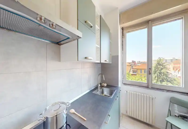 Compact, bright kitchen with a countertop, sink and large window offering city views — modern mint-green cabinets and neutral tiles provide a clean, airy feel.