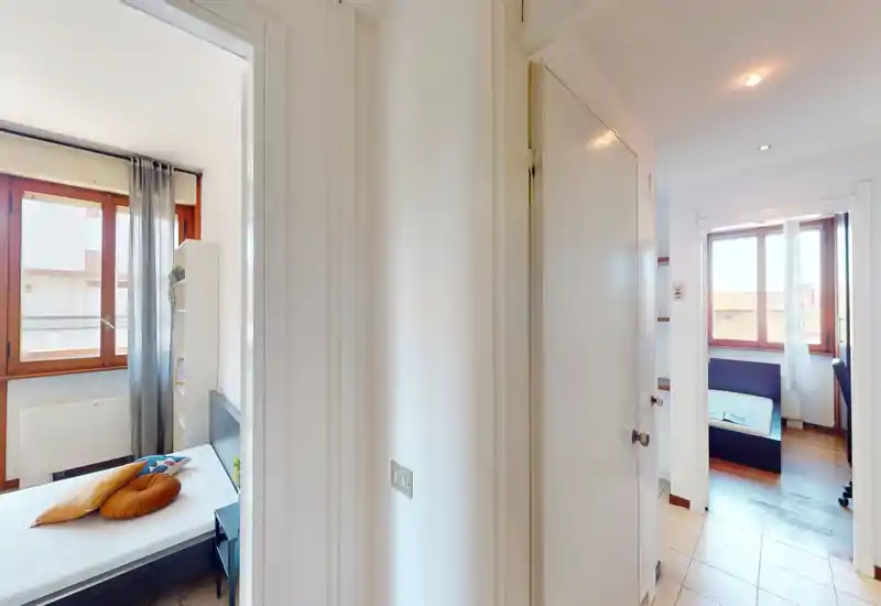 Immagine luminosa che mostra due piccole camere da letto viste da un corridoio centrale; in ogni stanza si vedono letto, cuscini e luce naturale dalle ampie finestre.