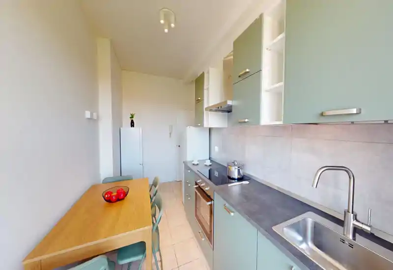 Bright, narrow galley kitchen with modern mint-green cabinets, integrated oven, sink and a wooden dining table with chairs — suitable for compact apartment listings.