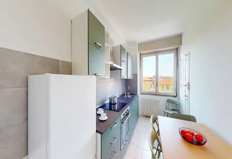 Bright, narrow kitchen with modern green cabinets, built-in oven and induction hob, a tall white fridge and a dining table by the window offering street views.