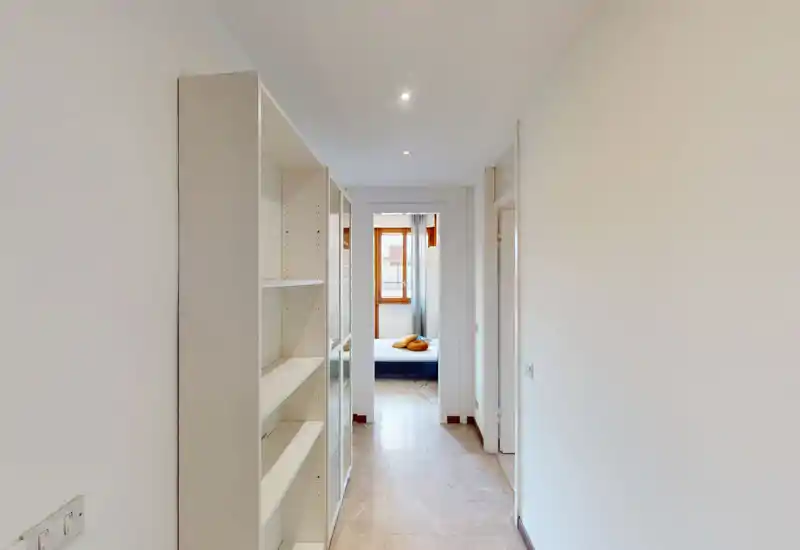 Narrow hallway view leading to a bedroom: you can see the bed through the doorway, built-in shelving along the corridor, and natural light from the bedroom window.