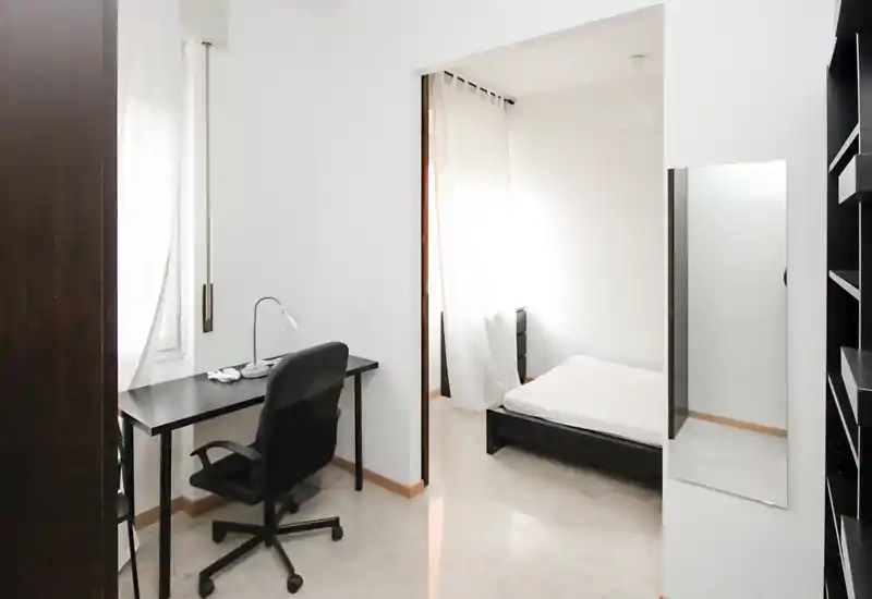 Camera da letto minimal e luminosa con struttura letto nera bassa e una scrivania semplice vicino alla finestra; pareti bianche e tende leggere creano un’atmosfera ariosa e pulita, ideale per affitti brevi.