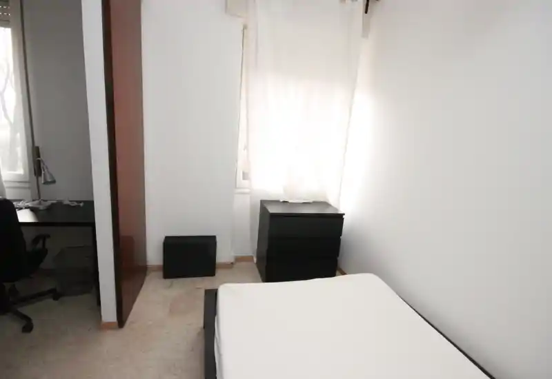 Camera da letto semplice e luminosa con letto singolo visibile, pareti bianche e comodini scuri; la luce naturale dalla finestra conferisce un aspetto pulito e minimale.