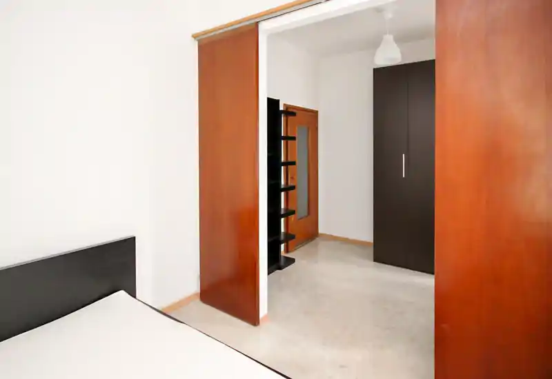 Angolo camera semplice con materasso e testata scura visibili, pareti bianche e porte scorrevoli in legno che conducono ad un'area adiacente; luminoso, minimale e funzionale.