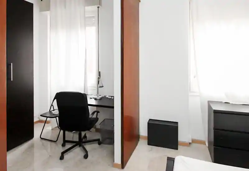 Piccola camera da letto con area studio: scrivania, sedia da ufficio e un angolo del letto visibili. La luce naturale attraverso tende leggere e mobili neri creano un ambiente minimale e funzionale.