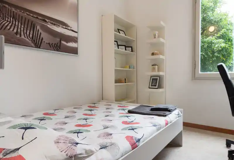 Dormitorio luminoso y ordenado con edredón estampado, estanterías abiertas y ventana con vistas a la vegetación: un espacio tranquilo y bien iluminado, ideal para estancias cortas.