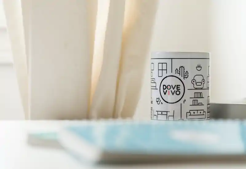 Primer plano de una taza con marca sobre una superficie blanca con cortinas suaves al fondo; poca profundidad de campo y un objeto desenfocado en primer plano.