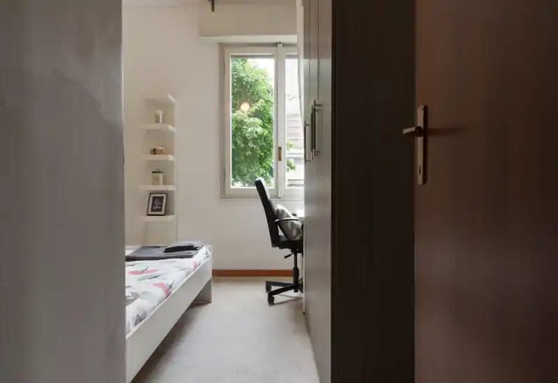 Dormitorio luminoso y sencillo con cama individual, estanterías y un escritorio junto a la ventana que ofrece luz natural y vistas al jardín.