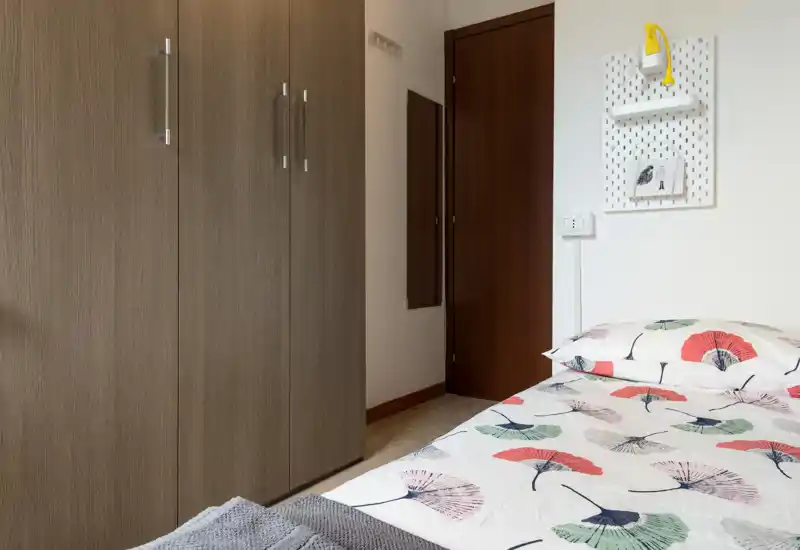 Rincón de dormitorio ordenado con edredón estampado, zona de la cama visible y armario empotrado — luminoso, limpio y con mobiliario sencillo.