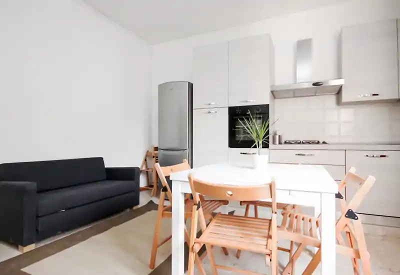Bright, minimalist open-plan living area with a small dining table, wooden folding chairs and a compact kitchen; a dark sofa provides seating and contrast.