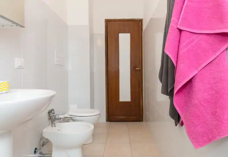 Clean, well-lit bathroom with a sink, bidet and toilet arranged along a tiled wall and a wooden door at the end; a pink towel hangs on the right.