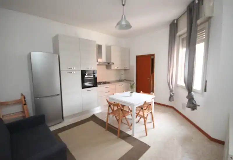 Bright open-plan kitchenette with integrated cabinets, oven, fridge and a small dining table for four; neutral décor and large windows provide a pleasant, airy feel.