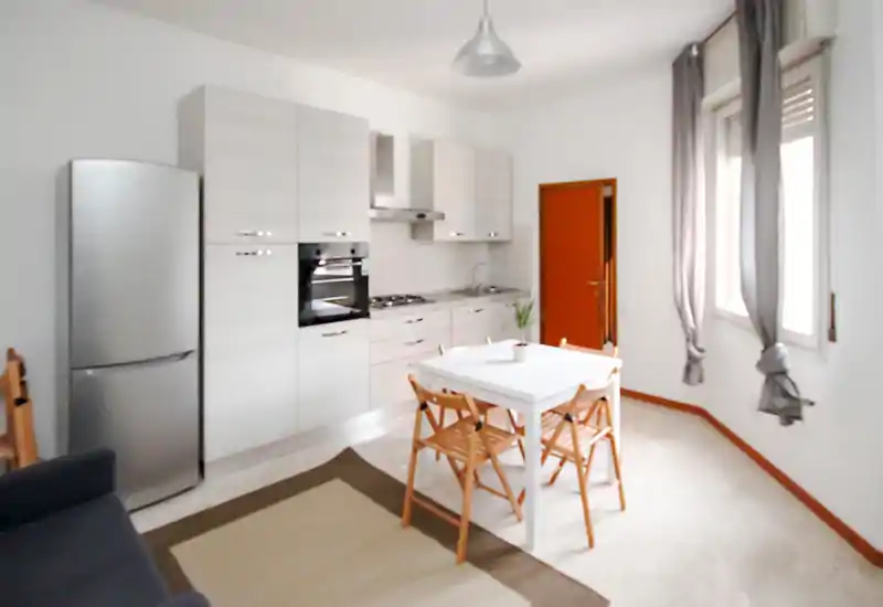 Bright compact kitchen with light cabinets, integrated oven and a small dining table. Natural light from the window and simple, cozy furnishings make this space welcoming for meals.