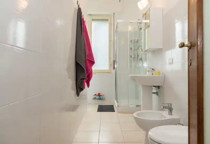 Clean, well-lit bathroom with walk-in shower, pedestal sink, bidet and toilet. Neutral tiles and bright window create an airy feel.