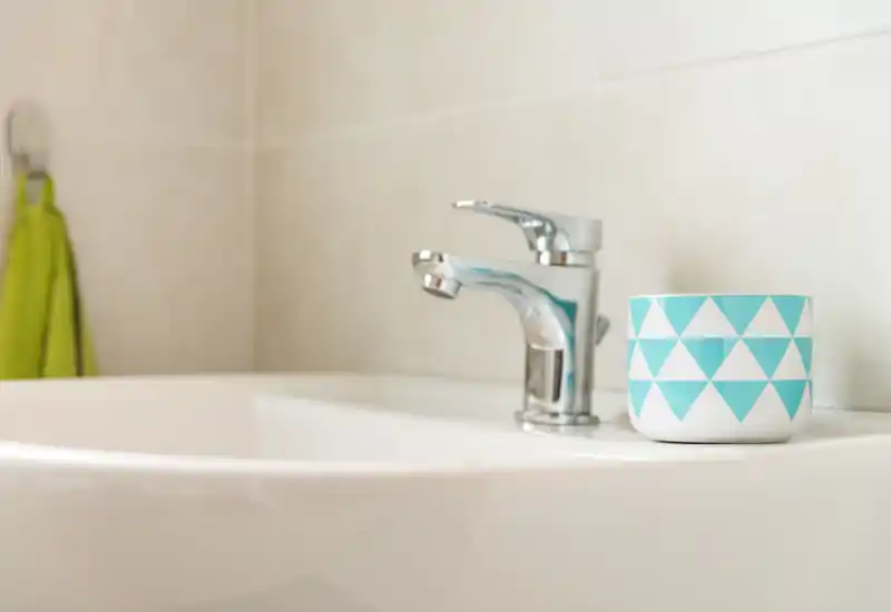 Close-up of a bathroom sink with a chrome faucet and a patterned cup on the basin, set against tiled wall — bright and clean detail shot ideal for listing bathroom fixtures.