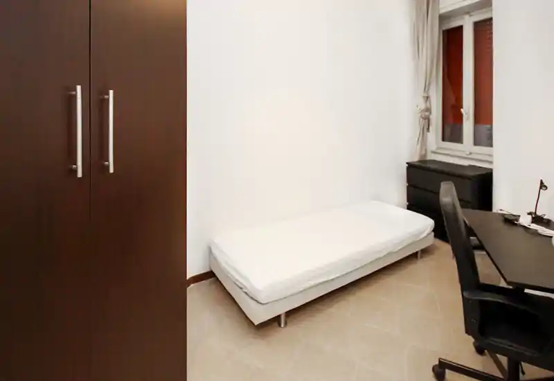 Chambre simple au style minimaliste : lit une place bien fait, armoire foncée, bureau et chaise près de la fenêtre. Espace lumineux et fonctionnel, idéal pour étudiant ou personne seule.