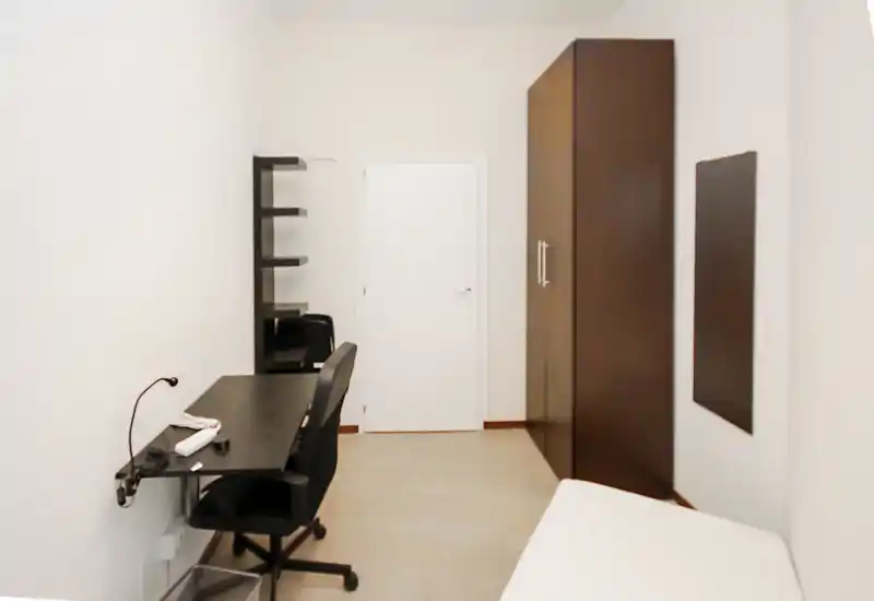 Chambre compacte avec bureau minimaliste et armoire. On aperçoit un coin de lit, un espace de travail avec chaise et étagères, et une grande armoire sombre dans une pièce claire et neutre.