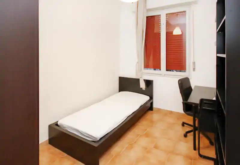 Camera singola semplice e ben illuminata con struttura letto bassa scura, biancheria bianca, scrittoio e sedia accanto alla finestra — ideale per studente o soggiorni brevi.