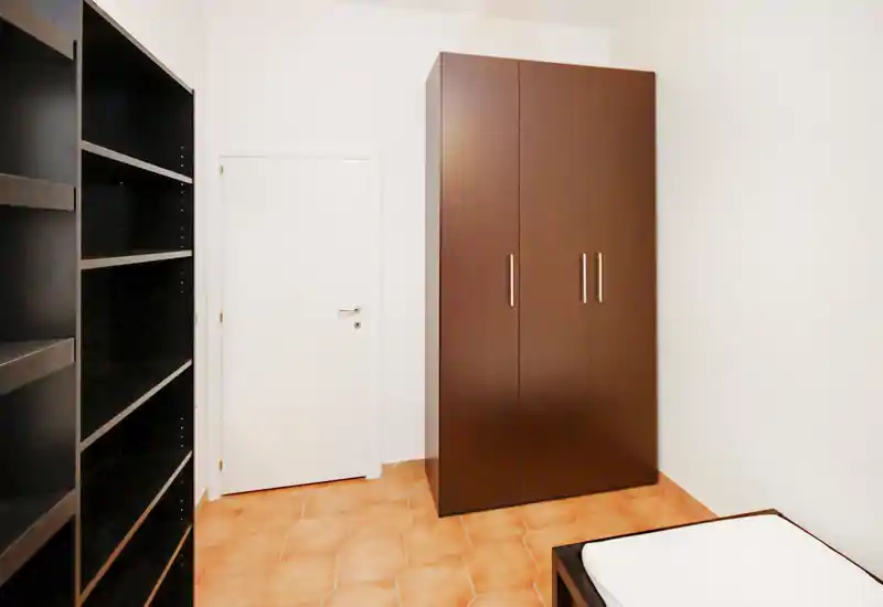 Angolo di camera da letto con armadio e scaffalatura visibili; nell'angolo anteriore si nota l'angolo di un materasso e una struttura letto semplice. Pareti bianche e pavimento in cotto creano un aspetto pulito e minimale.