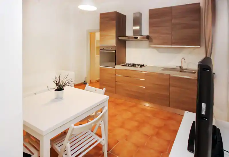 Cocina compacta y moderna con armarios efecto madera, placa de gas, horno integrado y fregadero de acero; suelo de baldosas cálido y una pequeña mesa que aportan una sensación acogedora y diáfana.