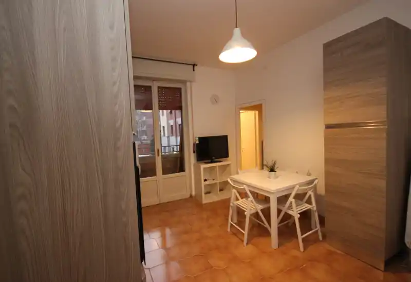 Pequeña zona de comedor con mesa blanca y cuatro sillas, lámpara colgante y mueble de TV; suelo de baldosas cálidas y puerta al balcón que aporta luz.