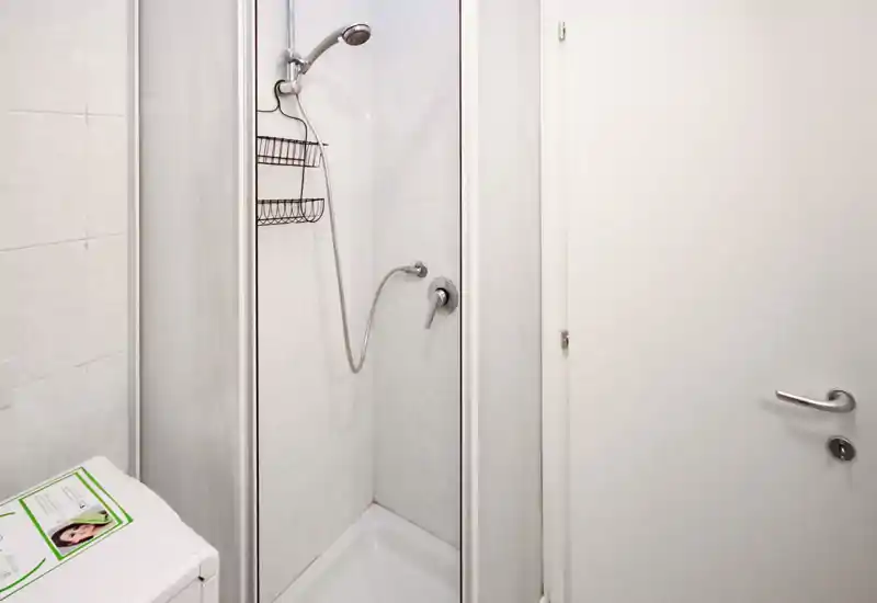 Baño compacto y limpio con ducha en esquina, alcachofa móvil y paredes alicatadas — luminoso, minimalista y funcional.