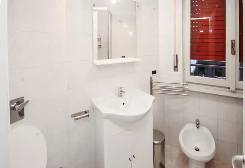 Baño compacto y limpio con mueble de lavabo, bidé y espejo de pared. Azulejos blancos y luz natural desde la ventana que ofrecen un espacio ordenado y práctico.