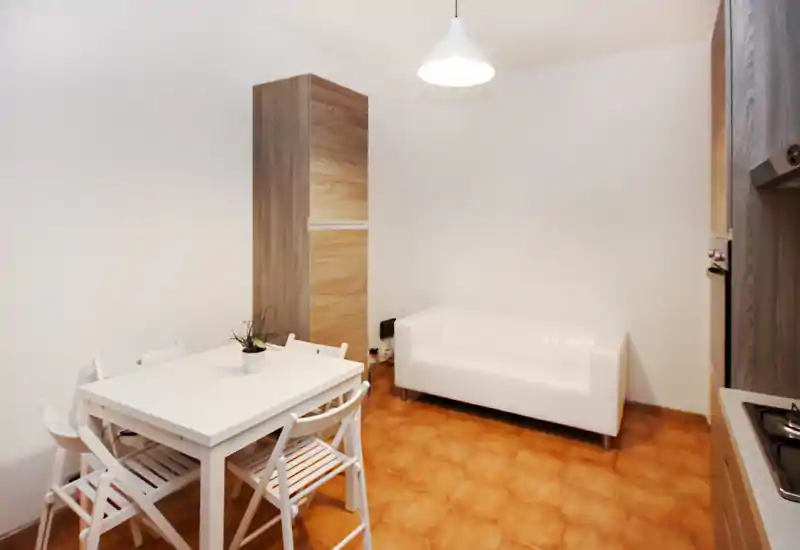 Espace de vie compact comprenant un petit canapé blanc et une table à manger avec chaises dans un intérieur lumineux et minimaliste. Le sol carrelé et les éléments boisés apportent une touche chaleureuse et fonctionnelle.
