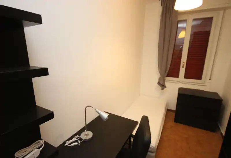 Habitación individual pequeña con colchón individual colocado, escritorio compacto y estanterías, cómoda y ventana con cortina que aporta luz natural.