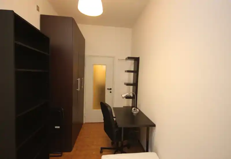 Piccola camera da letto con area studio: stanza stretta con angolo di letto singolo, scrivania con lampada e mensole, e un armadio alto. Pareti neutre e luce calda dall'alto creano uno spazio semplice e funzionale.