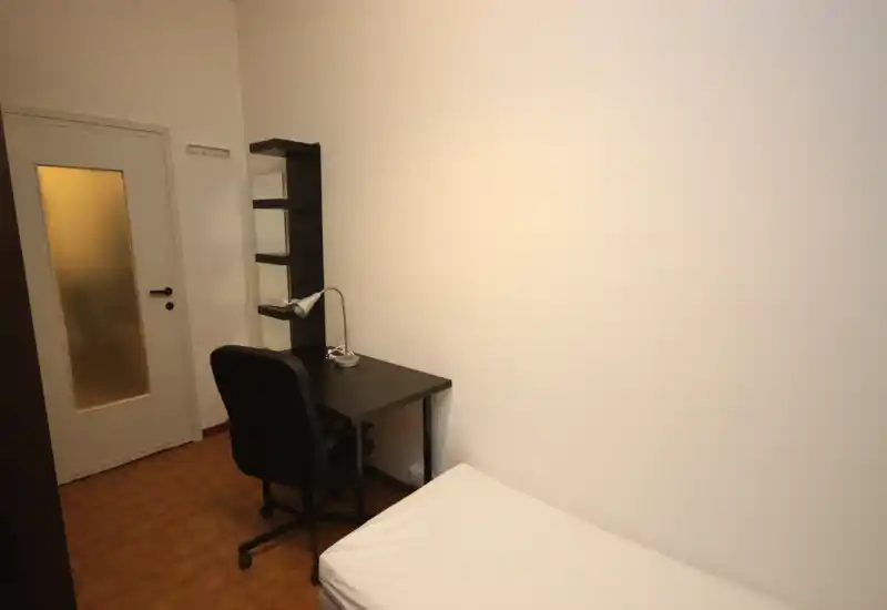 Camera compatta con letto singolo e scrivania semplice. Pareti neutre e pavimento caldo creano uno spazio funzionale e ordinato, ideale per studenti o soggiorni brevi.