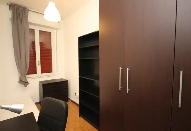 Camera da letto compatta con armadio in legno scuro, scaffalatura e un letto singolo parzialmente visibile a sinistra; pareti neutre, lampada da tavolo e finestra con tenda per illuminazione pratica.