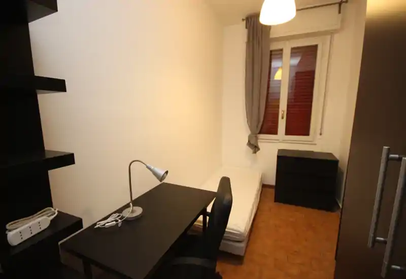 Dormitorio individual compacto con cama ordenada, escritorio y silla negros, cómoda y ventana que aporta luz natural; ideal para estudiante o ocupante individual.