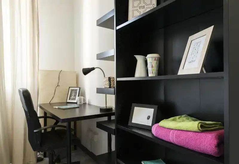 A tidy home office corner within a living area featuring a black desk, task chair and shelving with decorative frames and folded towels. Soft natural light from a nearby window creates a calm, functional workspace.