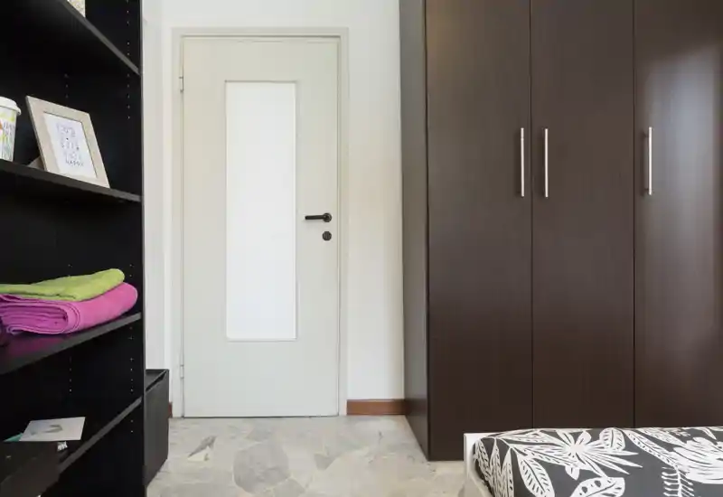 Coin de chambre avec un coin de lit visible, une grande armoire en bois foncé et des étagères ouvertes. L'espace est rangé, bien éclairé et met en évidence les solutions de rangement.