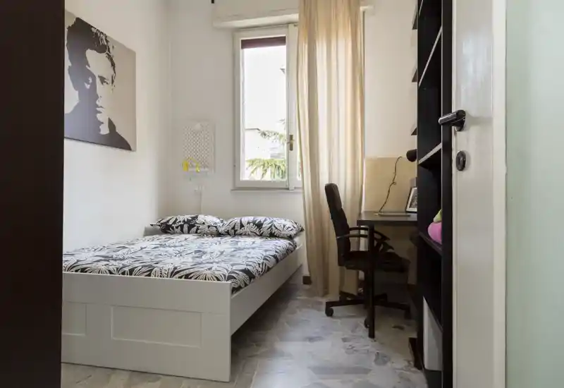 Chambre lumineuse et soignée avec un lit double, une parure à motifs et un petit espace de travail près de la fenêtre. La lumière naturelle et les tons neutres créent une ambiance confortable et bien éclairée.