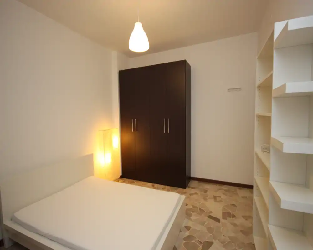 Camera da letto moderna e minimale con letto matrimoniale, armadio indipendente e scaffalatura aperta. Illuminazione calda da una lampada da terra e un lampadario, ambiente accogliente e ordinato ideale per il riposo.