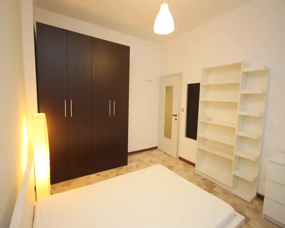 Camera da letto luminosa e arredata in modo semplice con letto matrimoniale in primo piano, armadio scuro e scaffalatura bianca. Lampada da terra calda e luce a soffitto creano un’atmosfera accogliente; pavimento a piastrelle e pareti bianche offrono un aspetto pulito e minimal.